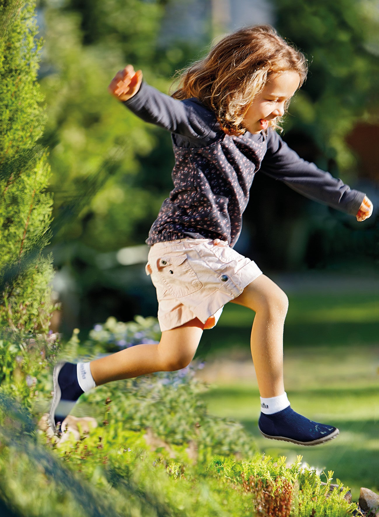 Leguanito Barfußschuhe für Kinder von Leguano in Blau – Sockenschuhe, getragen von einem Kind, das sich mitten im Sprung über eine grüne Hecke befindet, erhältlich im barfussweltan.shop Barfußschuhe Onlineshop