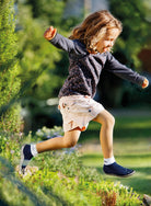 Leguanito Barfußschuhe für Kinder von Leguano in Blau – Sockenschuhe, getragen von einem Kind, das sich mitten im Sprung über eine grüne Hecke befindet, erhältlich im barfussweltan.shop Barfußschuhe Onlineshop